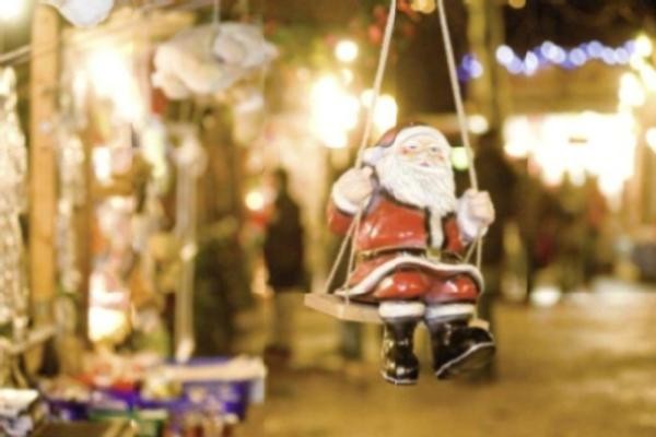 Weihnachtsmann aus Keramik auf einer Schaukel mit beleuchteten Weihnachtsmarktbuden im Hintergrund