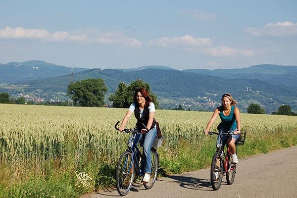 Zwei Fahrradfahrerinnen radeln auf einer Wirtschaftsstraße vor einem Weizenfeld und haben die Schwarzwaldberge im Hintergrund