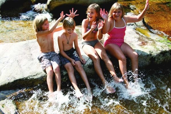 Vier Kinder sitzen auf einem großen Felsen und planschen mit den Füßem im Wasser