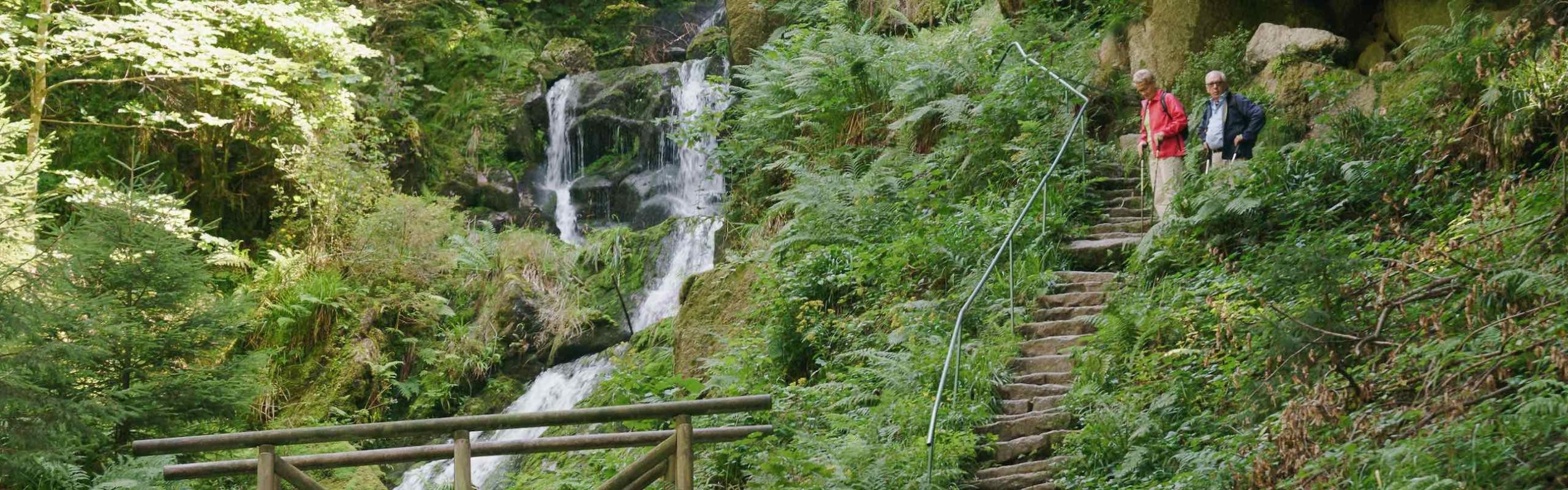 Gertelbach Wasserfälle in Bühlertal Das Bild zeigt zwei ältere Personen, die Steintreppen hochwanden und dabei Wasserfälle beobachten. Im unteren Bildbereich ist noch das Geländer einer kleinen Holzbrücke zu sehen