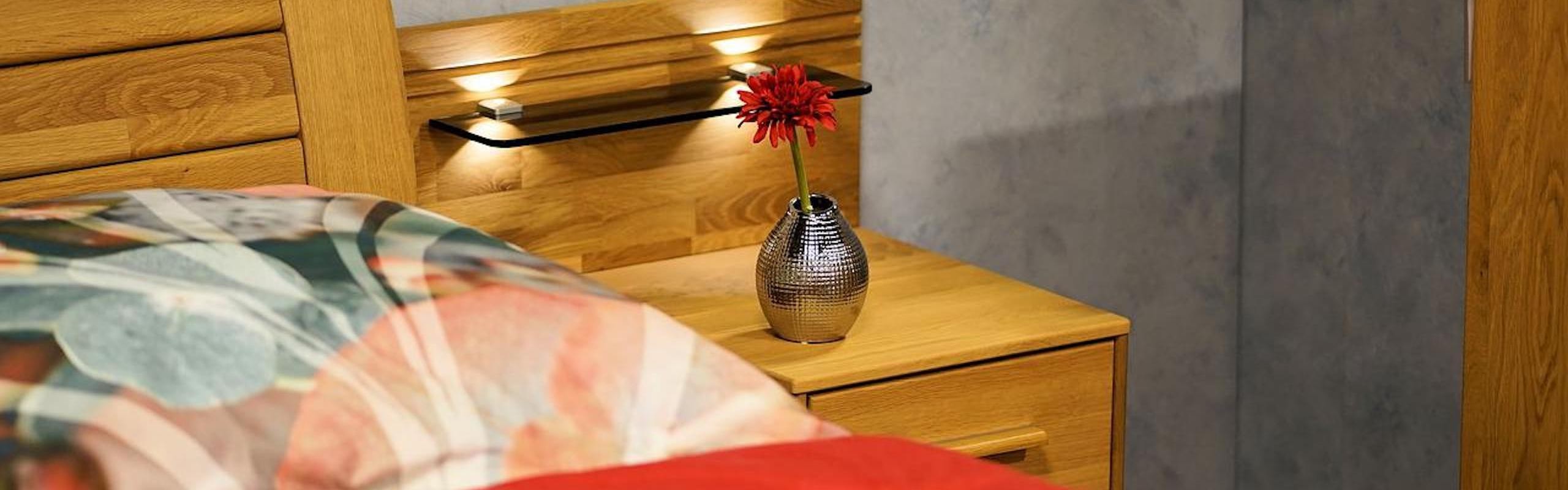 Ein gemütliches Holzbett mit bunter Bettwäsche und einer Blumenvase auf dem Nautisch mit einer roten Blume drin