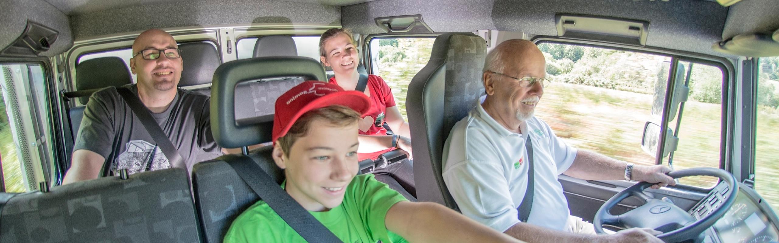 Das Bild zeigt vier Personen bei der Mitfahrt in einem Unimog. Ein Junge mit grünen T-Shirt und rotem Cap zeigt den Daumen hoch