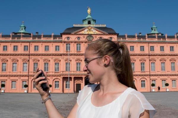 Das Bild zeigt eine Frau mit weißen T-Shirt & Brille und einem GPS-Gerät in der Hand vor dem Barockschloss Rastatt