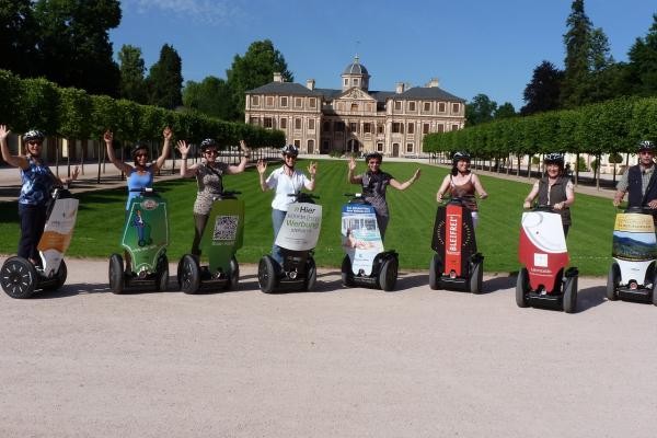 Das Bild zeigt acht winkende Personen stehend auf Segway vor dem Schloss Favoite
