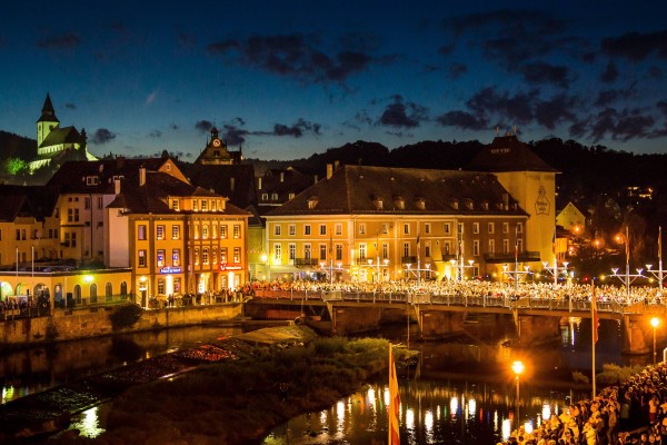 Das Bild zeigt zahlreiche Besucher in einer beleuchteten Altstadt auf einer Brücke und einer Flusspromenade am Abend 