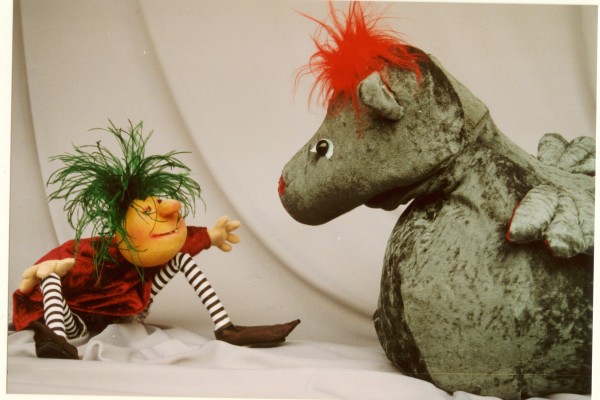 Das Bild zeigt einen Drachen und einen Menschen mit grünen, zotteligen Haaren als Puppentheaterfiguren vor weißem Hintergrund
