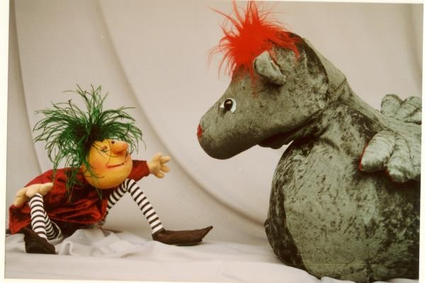 Das Bild zeigt zwei Puppentheaterfiguren. Einen grünen Drachen mit roten Haaren und einen sitzenden Menschen mit grünen, zotteligen Haaren 