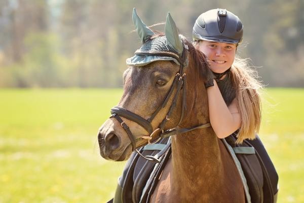 Das Bild zeigt ein grinsendes Mädchen mit langen blonden Haaren und Reithelm auf einem braunen Pferd