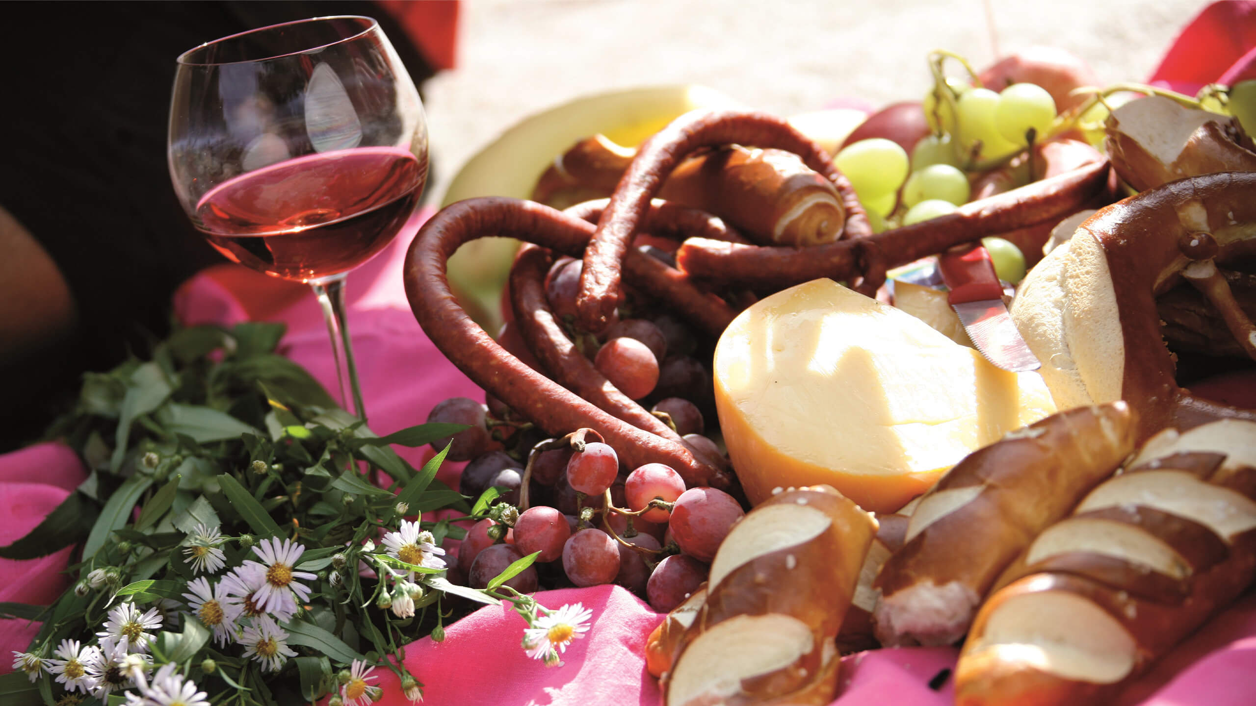 Typisches Vesper mit einem Glas Rotwein, Laugenstangen, Käse, Wurst und Weintrauben auf einer rosafarbenen Decke und Blumen als Dekoration